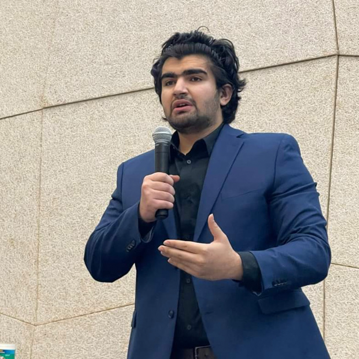 Ahmad wearing a blue suit and black shirt, speaking into a microphone